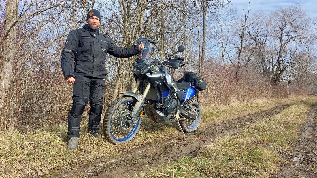 man standing beside his motorcycle wearing cold weather gryphon motorcycle jacket and gryphon motorcycle pants on a muddy wooded trail on a cold day