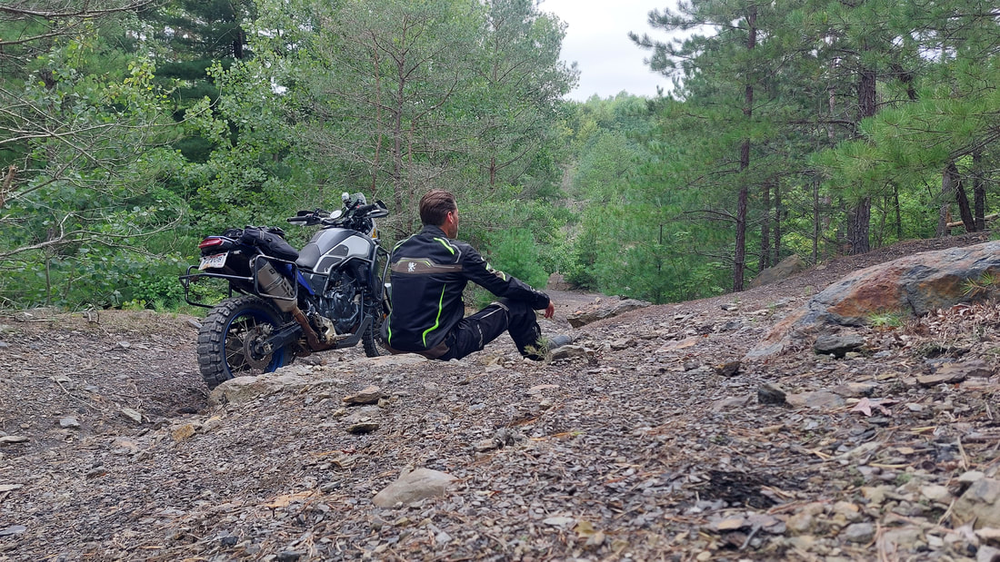 man sitting beside his motorcycle on a rocky wooded trail wearing gryphon motorcycle jacket and gryphon motorcycle pants 