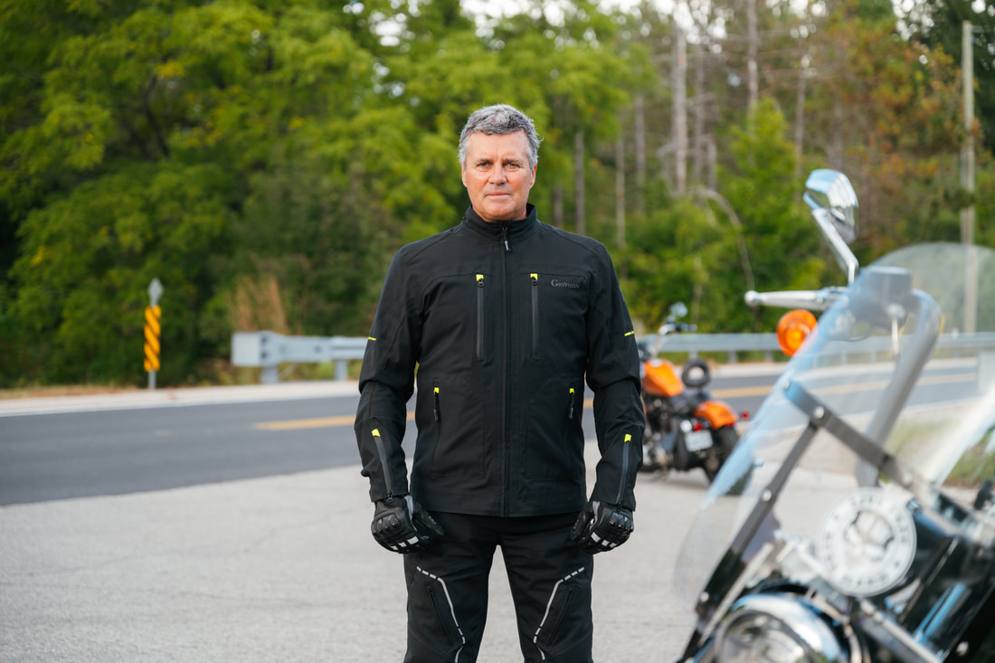 man standing on a road with a motorcycle in front and a motorcycle behind wearing gryphon motorcycle jacket and gryphon motorcycle pants and gryphon motorcycle gloves