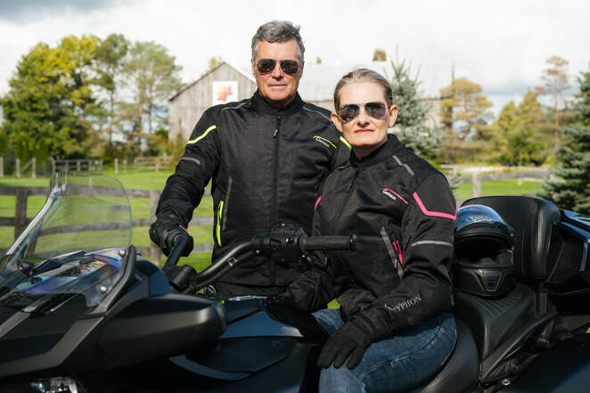 woman sitting on motorcycle with man standing behind wearing black Gryphon motorcycle jackets and sunglasses