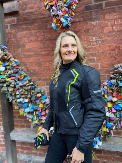 woman wearing gryphon motorcycle jacket and holding a pair of gryphon motorcycle gloves smiling in front of a brick wall with a sculpture 