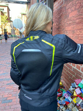woman standing beside a brick wall facing away wearing a gryphon motorcycle jacket 