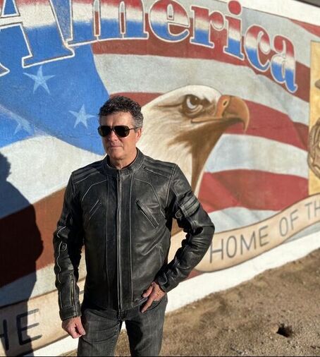man wearing a leather gryphon motorcycle jacket standing beside a large american flag mural with eagle 