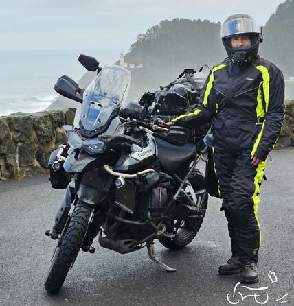 a woman standing beside her motorcycle wearing a gryphon rain suit with the ocean and mountains in the background on a cloudy and rainy day