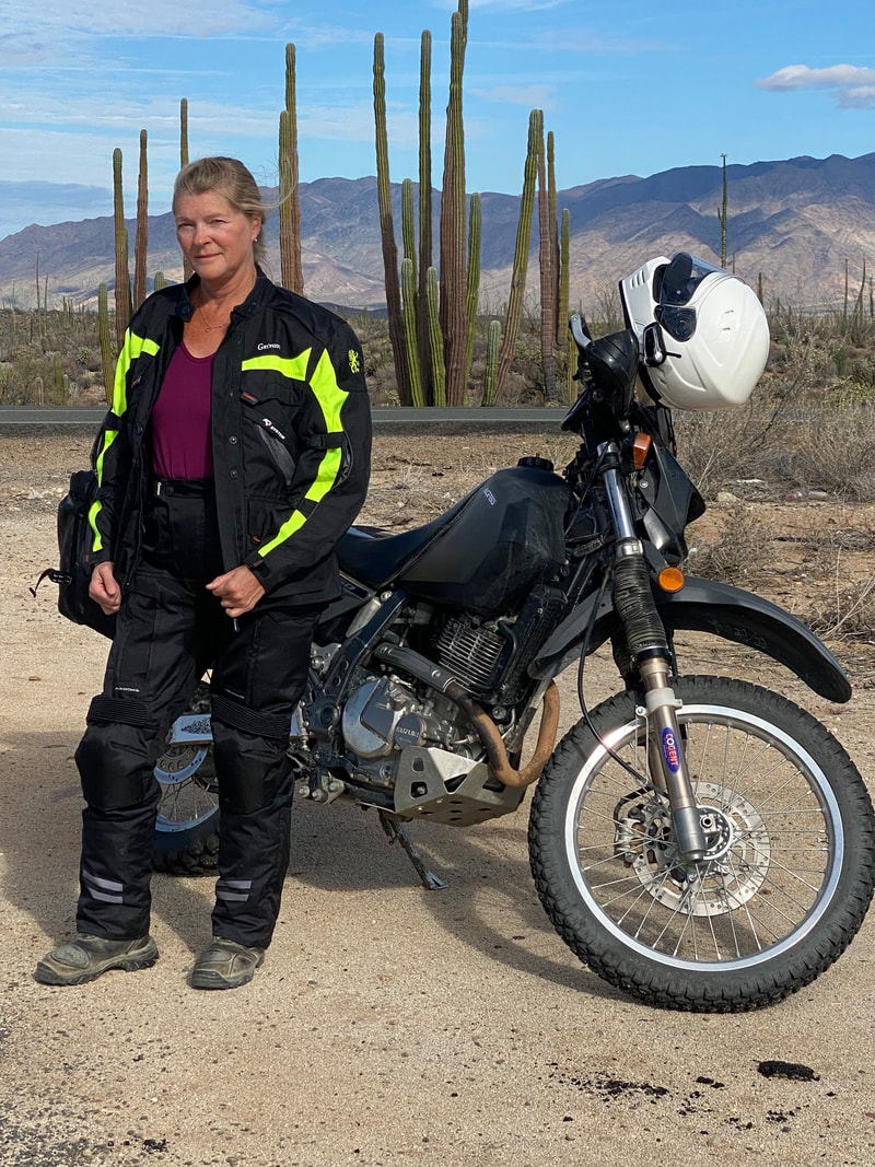 woman standing infront of her motorcycle in the desert with cacti in the background wearing gryphon motorcycle jacket and gryphon motorcycle pants 